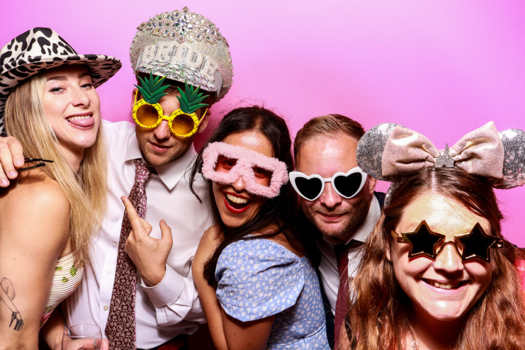 A group of five people wearing colourful and quirky hats and glasses, posing together in front of a bubblegum pink photo booth backdrop. They are smiling and making playful gestures towards the camera.