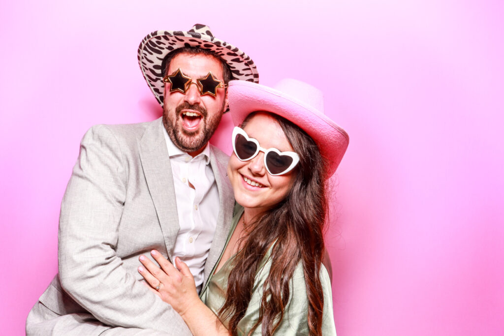 A couple wearing star-shaped and heart-shaped sunglasses, with the man also wearing a leopard print hat and the woman a pink cowboy hat, posing happily in front of a bubblegum pink photo booth backdrop.