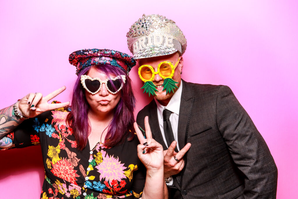 A woman with purple hair and a man in a suit posing in front of a bubblegum pink photo booth backdrop. They are both wearing novelty hats and glasses, making peace signs towards the camera.