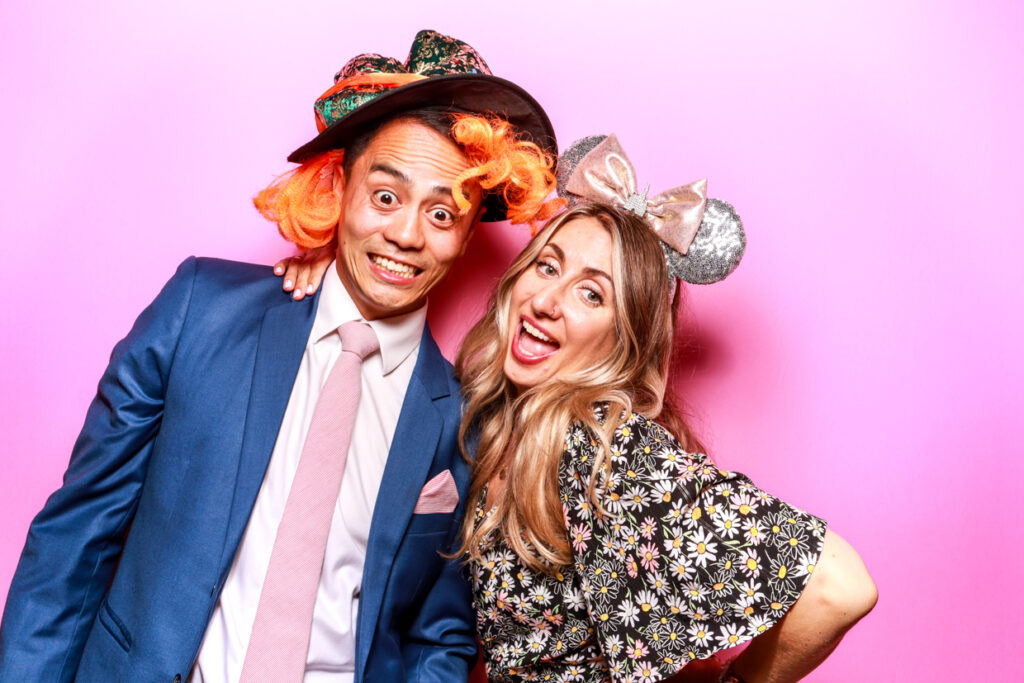 A man in a blue suit and a woman in a floral dress posing together in front of a bubblegum pink photo booth backdrop. The man is wearing an orange wig and a large hat, while the woman wears a large bow headband, both smiling widely.