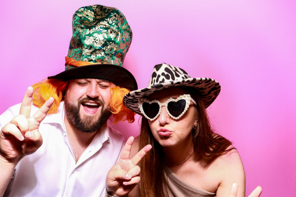 A man and a woman wearing novelty hats and glasses posing in front of a bubblegum pink photo booth backdrop. The man is wearing a tall, green hat with orange side curls, and the woman wears a leopard print hat and heart-shaped sunglasses, both making peace signs.