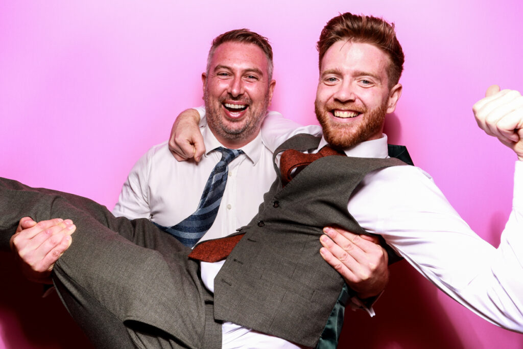 Two men in formal attire posing in front of a bubblegum pink photo booth backdrop. One man is joyfully holding the other in his arms, both smiling widely at the camera, creating a fun and lively atmosphere.