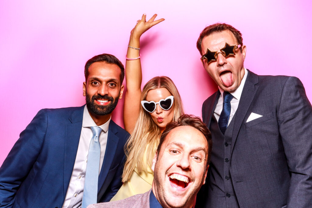  A group of four people dressed in formal attire, posing in front of a bubblegum pink photo booth backdrop. One man is smiling widely in the foreground, another man in a blue suit is smiling next to a woman wearing heart-shaped sunglasses, and a man in star-shaped sunglasses is playfully sticking out his tongue.