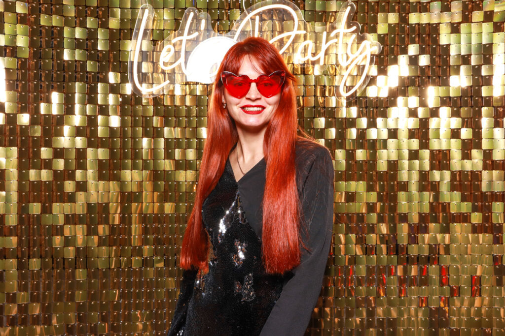 A woman with long red hair wearing red sunglasses and a black outfit smiles while standing in front of a gold shimmer backdrop with a "Let's Party" neon sign at estelle manor venue