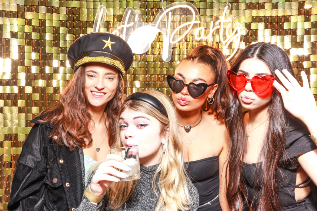 A group of four women, two wearing heart-shaped sunglasses and one in a captain's hat, pose together in front of a gold shimmer backdrop with a "Let's Party" neon sign. One woman is sipping a drink, and the others are smiling and making playful expressions.