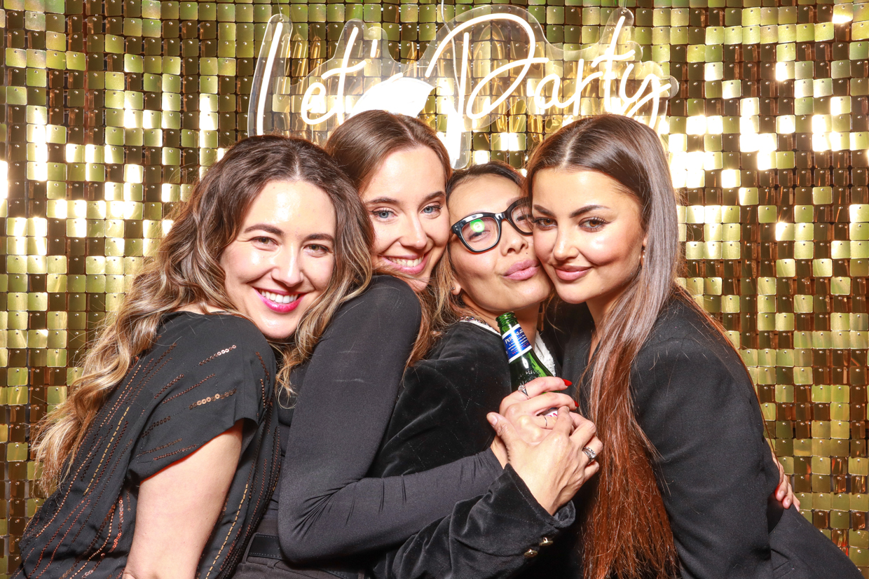 A group of four women dressed in black hug and smile for the camera in front of a gold shimmer backdrop with a "Let's Party" neon sign. One woman is holding a drink bottle, and they all appear to be enjoying the moment at a corporate event at Estelle manor