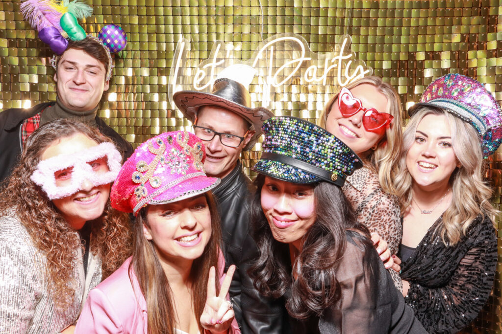 A group of seven people wearing colourful hats and playful accessories pose together in front of a gold shimmer backdrop with a "Let's Party" neon sign. They are smiling, making funny faces, and showing peace signs, enjoying the moment at a corporate event.