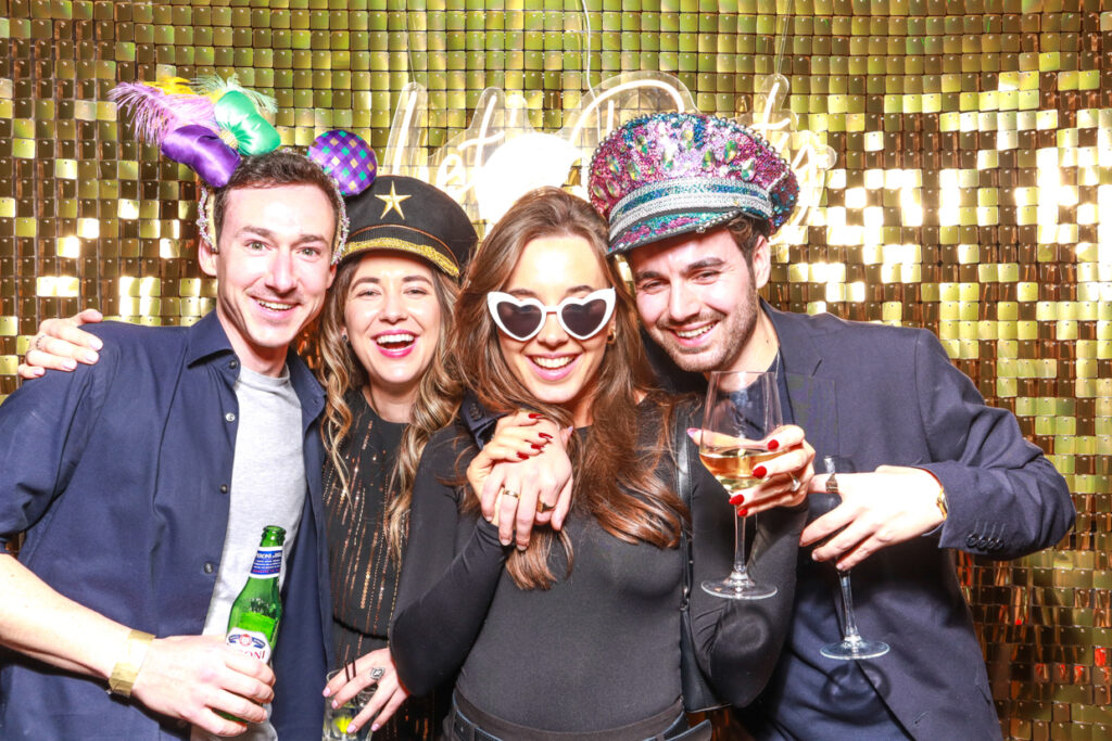 A group of four people wearing fun hats and accessories pose together, smiling and holding drinks, in front of a gold shimmer backdrop with a "Let's Party" neon sign