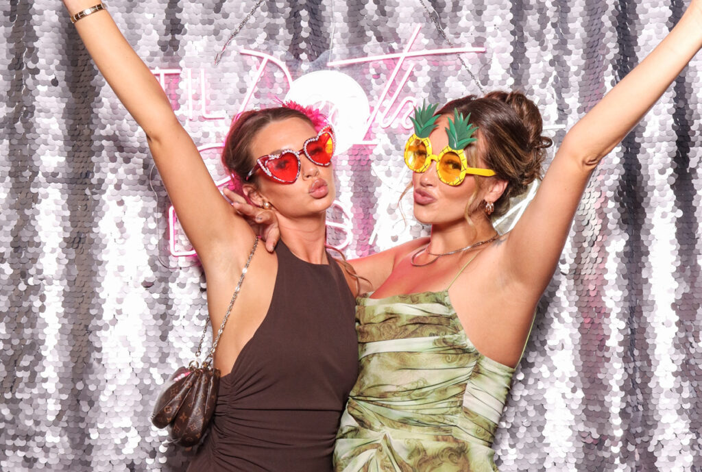 Two women wearing heart-shaped and pineapple-shaped sunglasses posing enthusiastically in front of a silver sequins backdrop with a neon sign reading "Till Death Do Us Party.