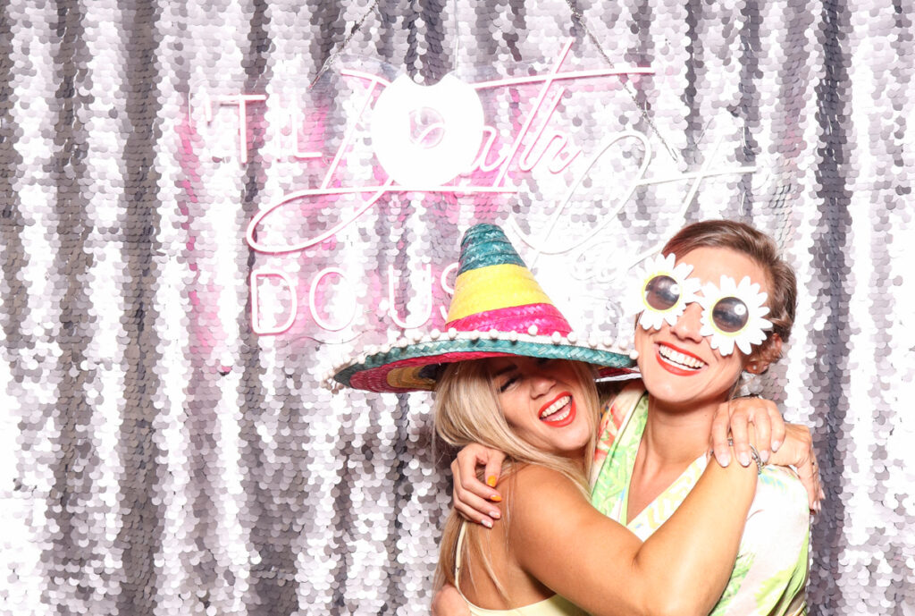 two women posing happily in front of a silver sequins backdrop with a neon sign reading "Till Death Do Us Party for a Wedding at Hampton Manor