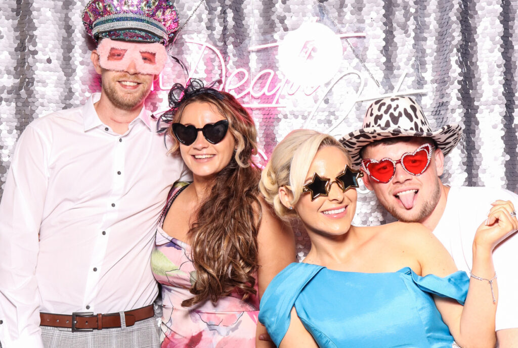 A group of four people, including two women and two men, all wearing playful sunglasses and hats, posing happily in front of a silver sequins backdrop with a neon sign reading "Till Death Do Us Party for a Wedding at Hampton Manor