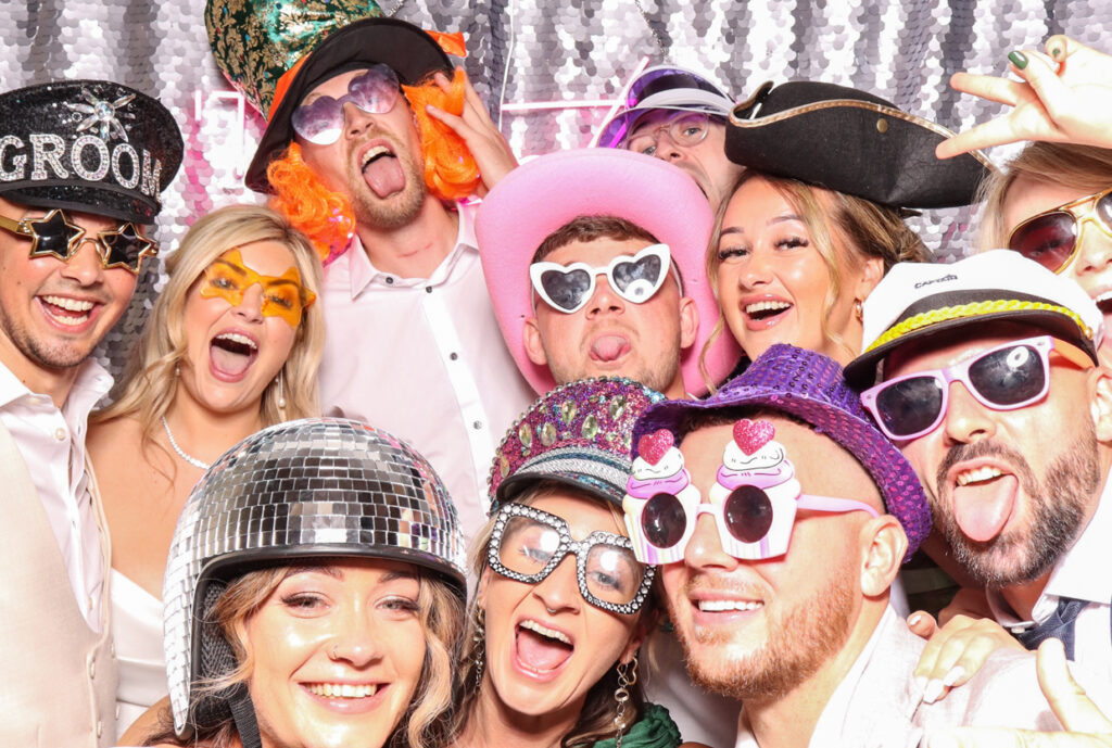 A large group of wedding guests wearing an assortment of funny sunglasses and hats, all smiling and posing energetically in front of a silver sequins backdrop with a neon sign reading "Till Death Do Us Party.