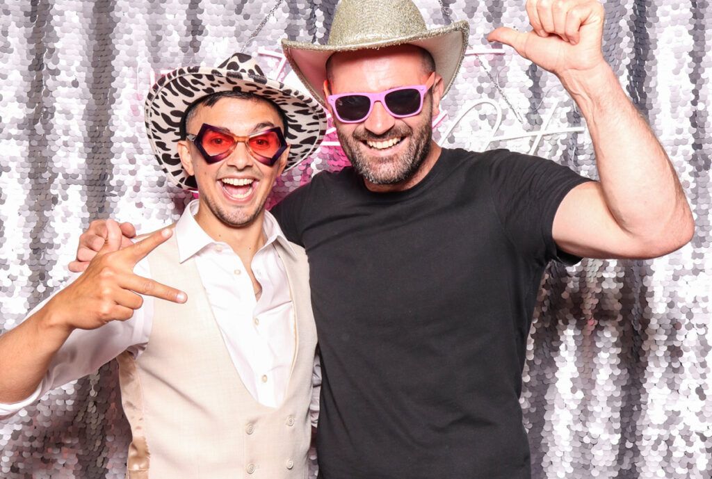 Two men, one in a vest and the other in a black shirt, both wearing sunglasses and hats, smiling and posing in front of a silver sequins backdrop with a neon sign reading "Till Death Do Us Party.