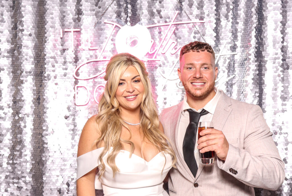 the bride and groom smiling and posing in front of a silver sequins backdrop with a neon sign reading "Till Death Do Us Party.