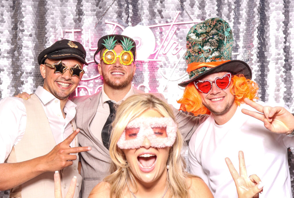 A group of four people, including two women and two men, all wearing playful sunglasses and hats, posing happily in front of a silver sequins backdrop with a neon sign reading "Till Death Do Us Party for a Wedding at Hampton Manor