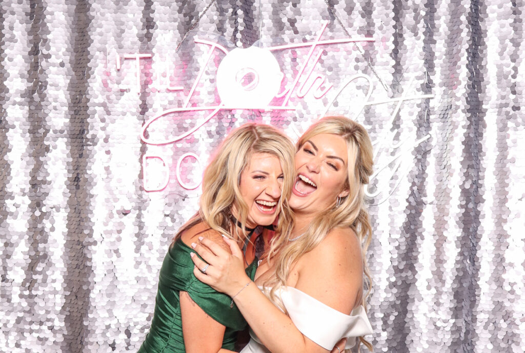 two women posing happily in front of a silver sequins backdrop with a neon sign reading "Till Death Do Us Party for a Wedding at Hampton Manor