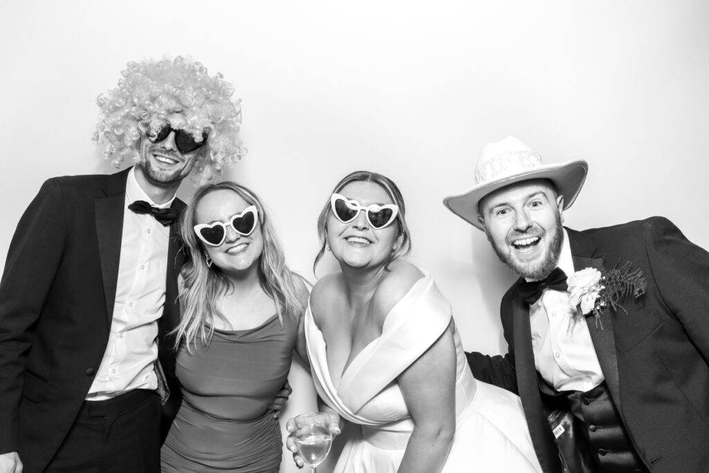 Four wedding guests posing with fun props in a photo booth at Photo Booth at Manor Hill House. They are wearing heart-shaped sunglasses and hats, smiling and enjoying the moment