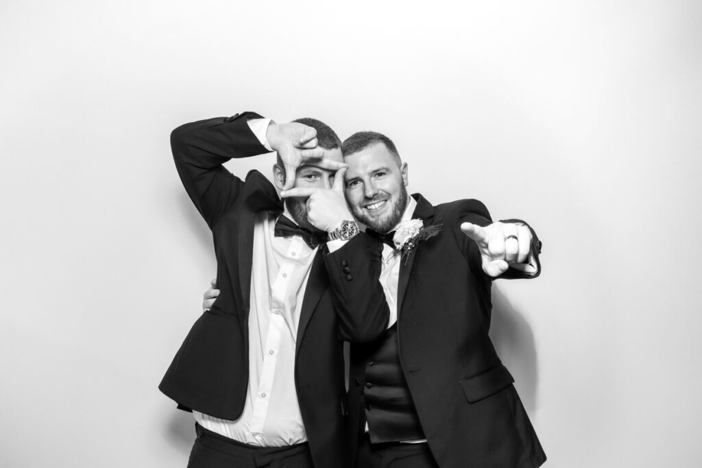 Two wedding guests in suits posing playfully in a photo booth. One is framing his face with his hands, while the other points towards the camera