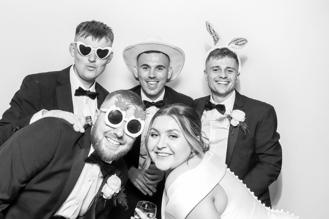 Five wedding guests, including the bride, wearing various props such as sunglasses and hats, crowded together and smiling in the photo booth at manor hill house
