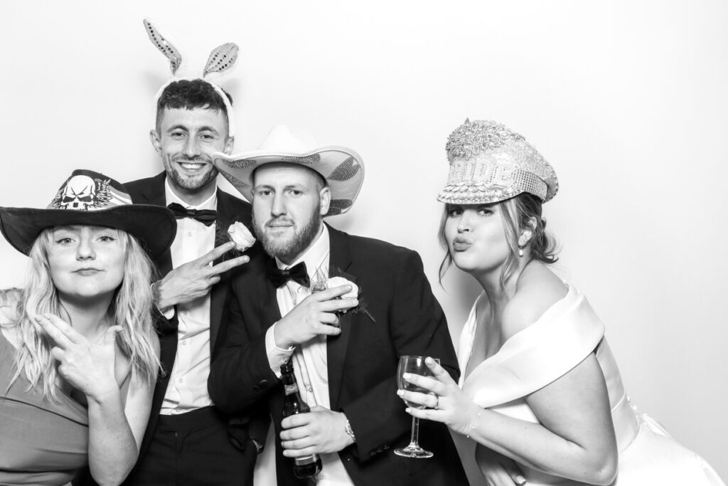 Four wedding guests, including the bride, posing with cowboy hats and bunny ears in a photo booth. They are holding drinks and making peace signs