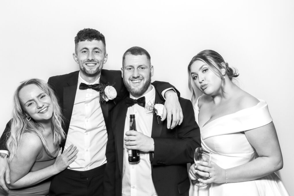 Four wedding guests, including the bride and groom, posing happily in a photo booth. They are smiling and holding drinks, enjoying the celebration