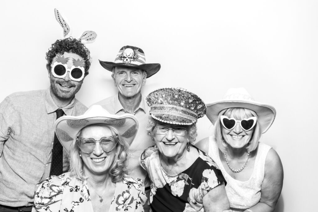 A group of five older wedding guests, including two men and three women, posing with fun hats and sunglasses in a photo booth at Manor Hill House, all smiling warmly.