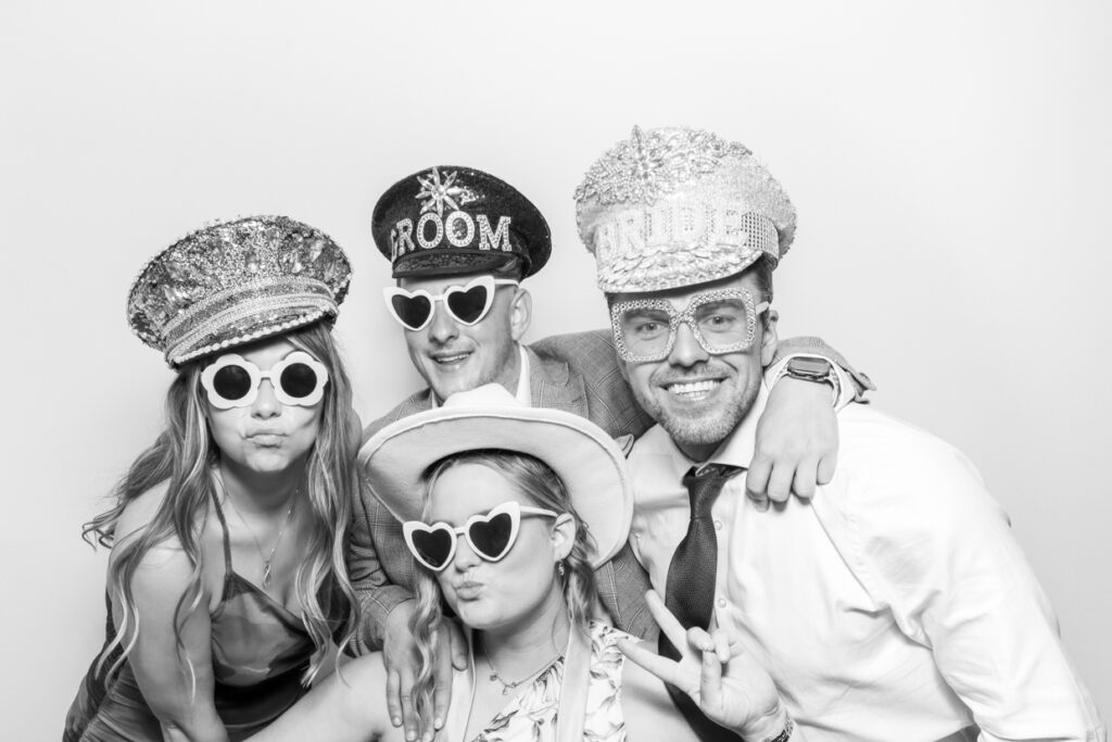 Four wedding guests wearing festive hats and heart-shaped sunglasses, posing together and making playful gestures in the Photo Booth at Manor Hill House