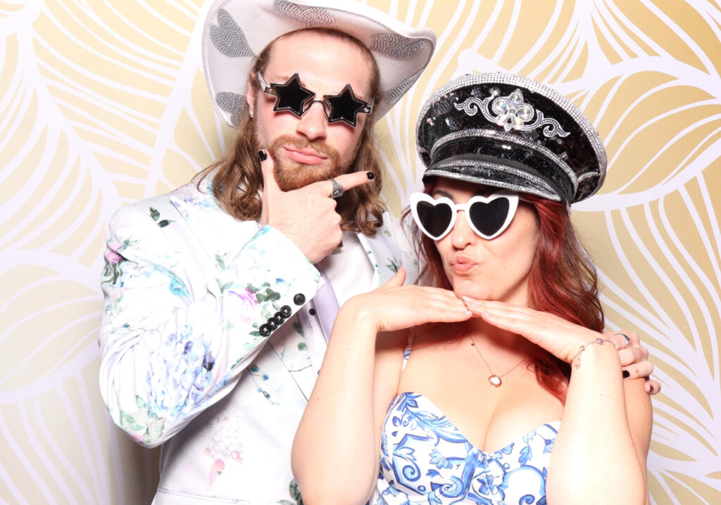 A man wearing star-shaped sunglasses and a white cowboy hat poses with a woman wearing heart-shaped sunglasses and a sparkly captain's hat. Both are making playful expressions and gestures.