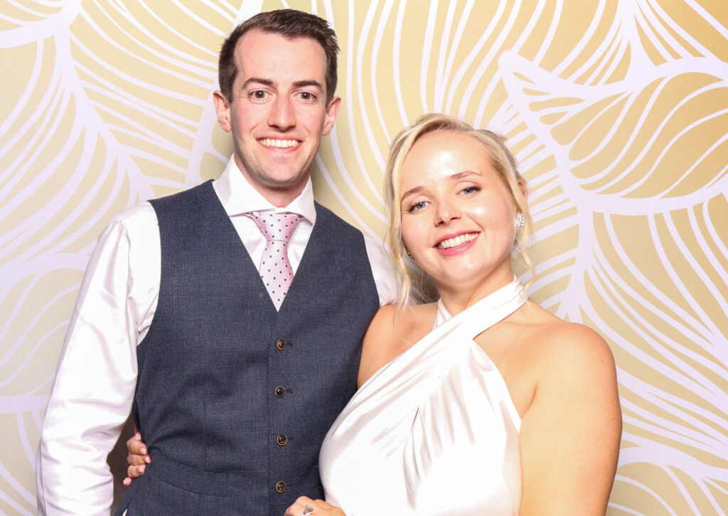 A smiling couple poses together in front of a patterned backdrop. The man is wearing a grey waistcoat, white shirt, and pink tie, while the woman is dressed in a white halter neck dress. Both are looking directly at the camera with happy expressions