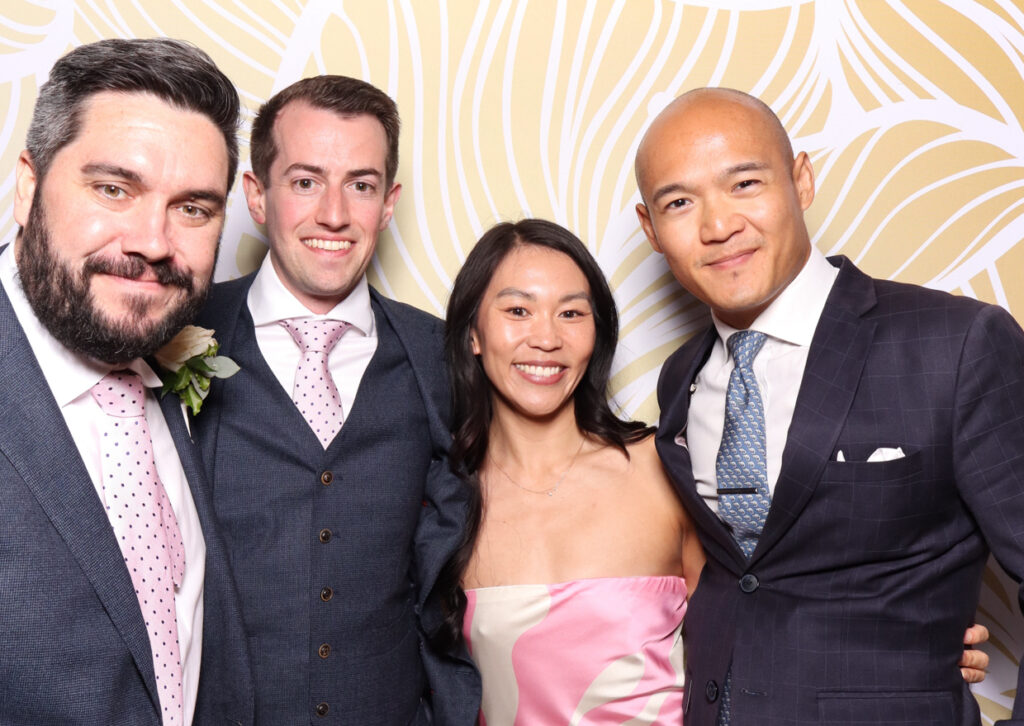 A group of four friends dressed in formal attire, smiling at the camera. The men are wearing suits and ties, while the woman in the centre is in a pink strapless dress. They look happy and close, enjoying the wedding celebration