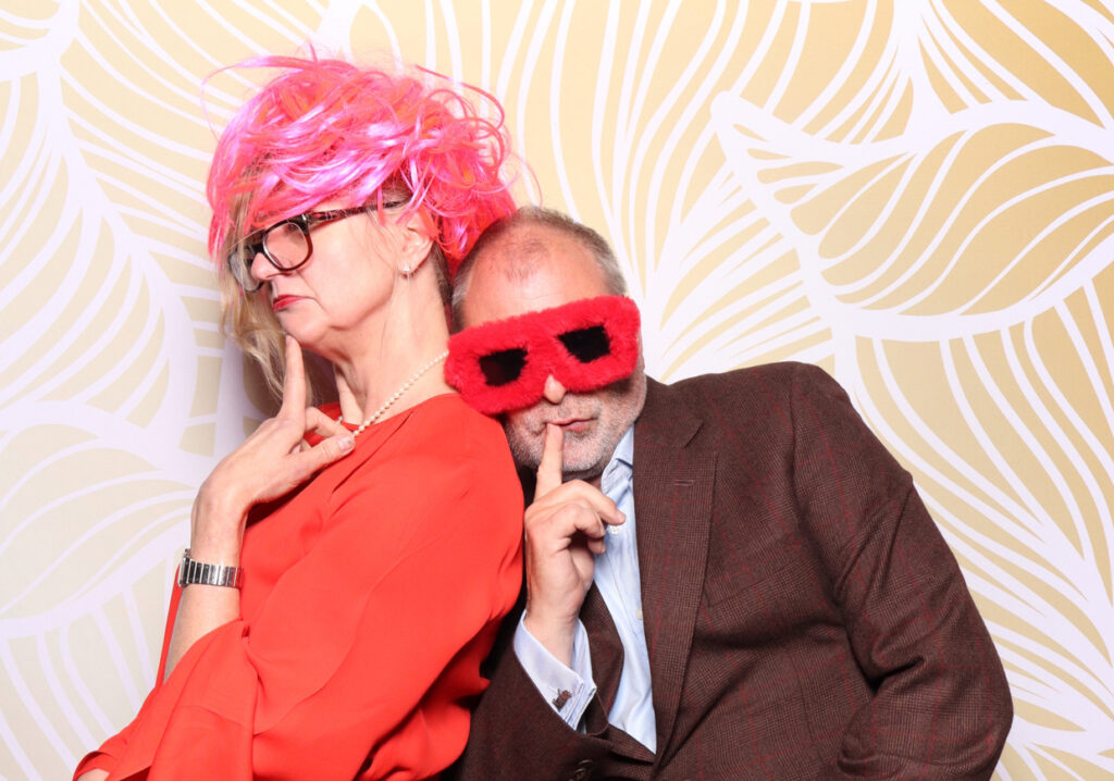 An older couple posing playfully. The woman is wearing a bright pink wig and glasses, looking serious, while the man has on fuzzy red sunglasses and is making a shushing gesture with his finger to his lips. Both are dressed in formal attire, adding a humorous touch to the photo