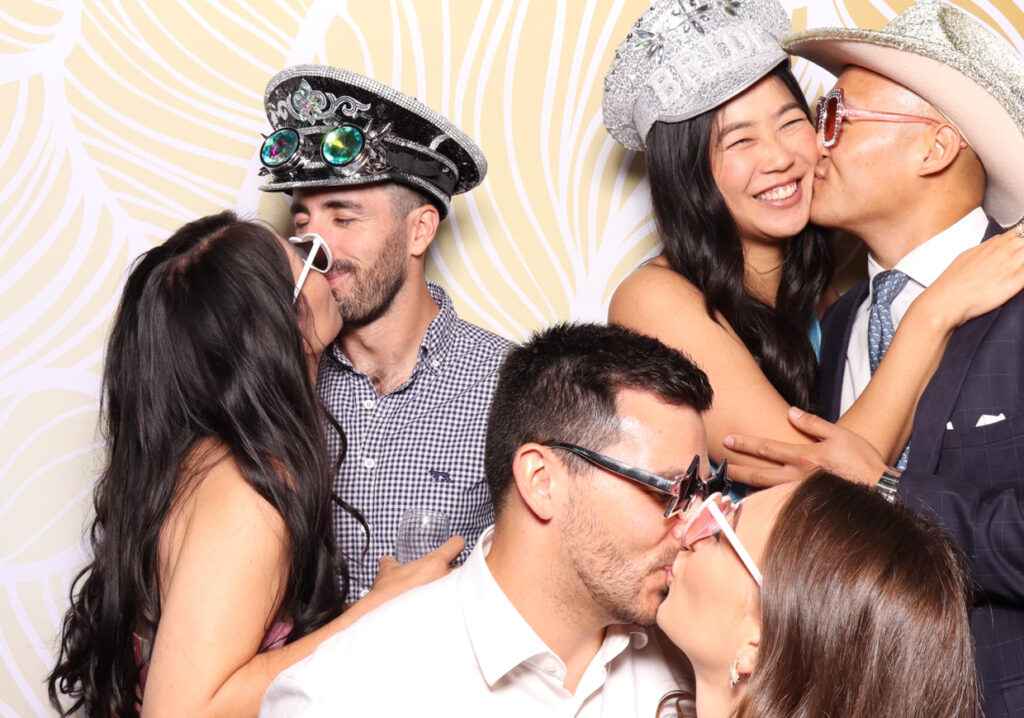 Three couples, each wearing fun hats and sunglasses, share kisses. The couples are dressed in a mix of formal and playful attire, enjoying a moment of affection and laughter in the photo booth for a Sudeley Castle Wedding 