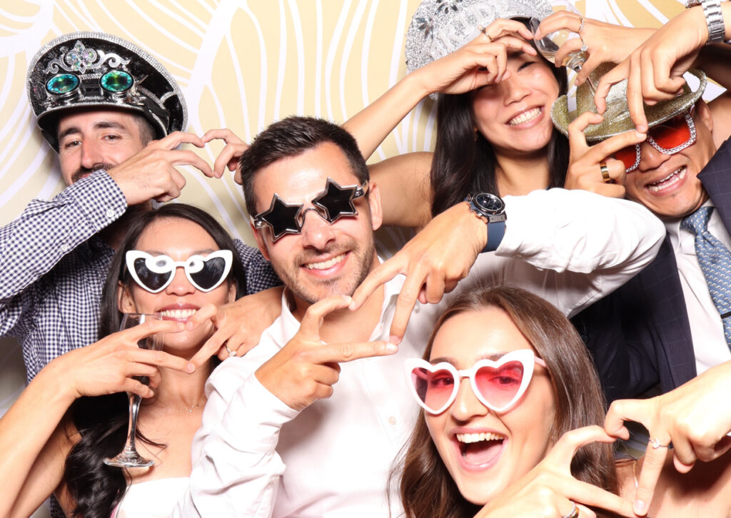 A group of friends, all wearing various quirky sunglasses and hats, forming heart shapes with their hands and smiling broadly at the camera. They are clearly enjoying the fun of the photo booth.