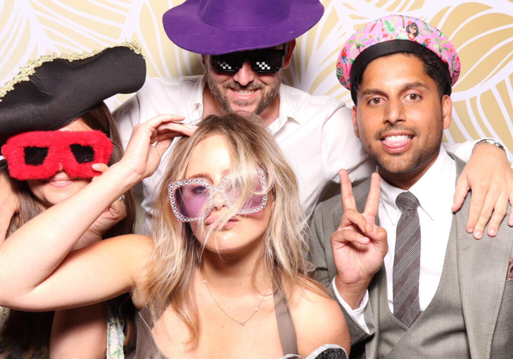 Four people, dressed in fun and quirky hats and sunglasses, pose together. One person wears a black pirate hat, another a purple hat, and all are making playful gestures and expressions.