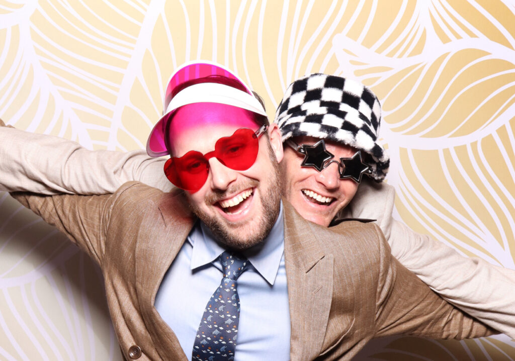 Two men with big smiles, each wearing playful hats and sunglasses. One wears a pink visor and heart-shaped glasses, while the other wears a checkered hat and star-shaped glasses. They have their arms around each other for a Sudeley Castle Wedding 