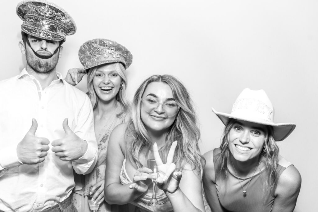 Four guests posing in a photo booth with fun hats and props, giving thumbs up and peace signs, creating a lively and memorable moment."