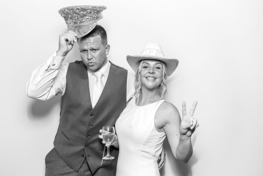 Bride and groom in the wedding photo booth, the bride with arms outstretched and the groom giving thumbs up, both smiling widely