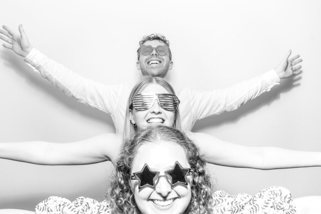 Three wedding guests in a photo booth, wearing fun hats and sunglasses, with one guest striking a playful pose with arms raised and tongue out.