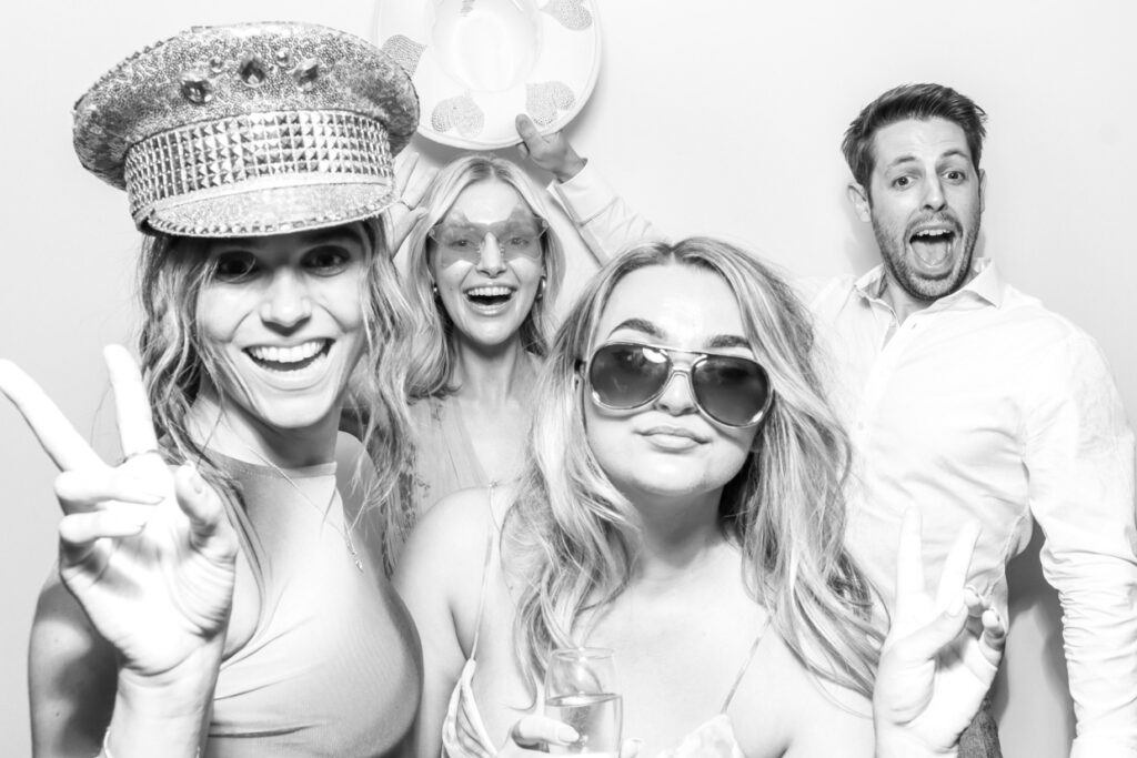 Four guests posing in a photo booth with fun hats and props, giving thumbs up and peace signs, creating a lively and memorable moment.