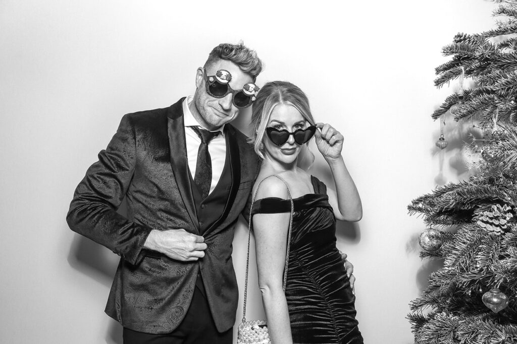 couple dressed in elegant festive outfits during a christmas party event, posing against a white backdrop with a black and white filter