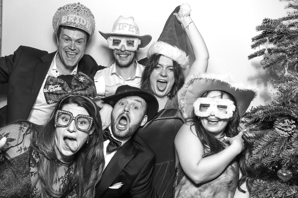 a large group of guests posing with christmas props and christmas hats, against a white backdrop with a christmas tree on one side, posing for mad hat photo booth at old gore by yard events in the cotswolds