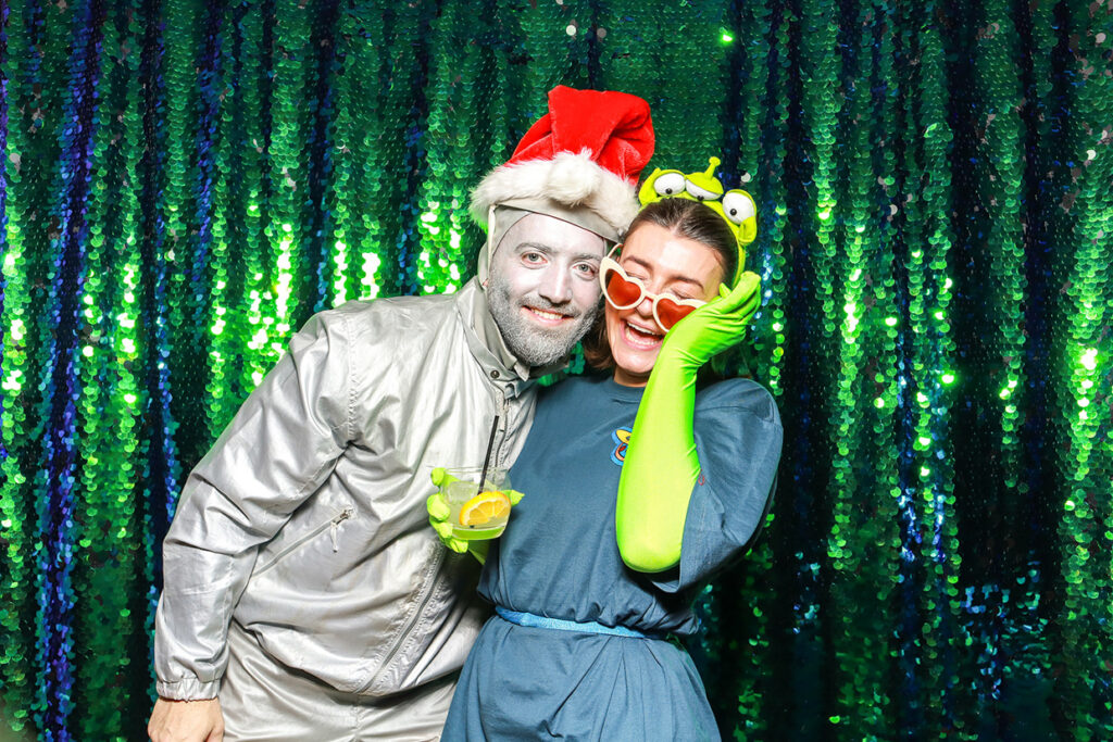 a couple wearing christmas hats posing against a green sequins backdrop, during a festive photo booth rentals company for corporate events and staff parties