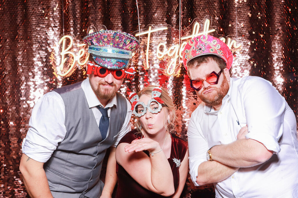 3 guests wearing hats and Christmas props posing against a champagne sequins backdrop during a photo booth party rental with real-time social media sharing