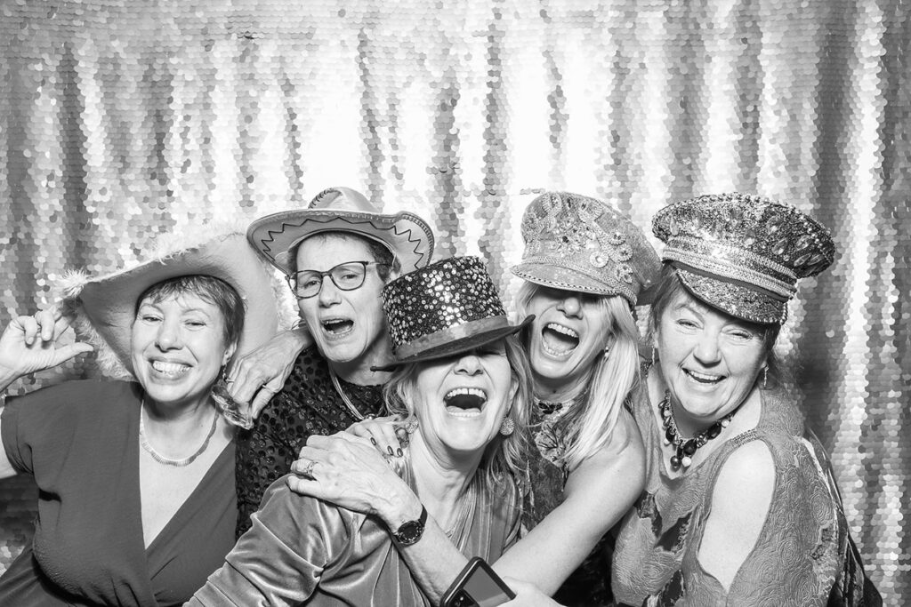 5 ladies wearing hats and Christmas props posing against a silver sequins backdrop during a photo booth party rental with real-time social media sharing