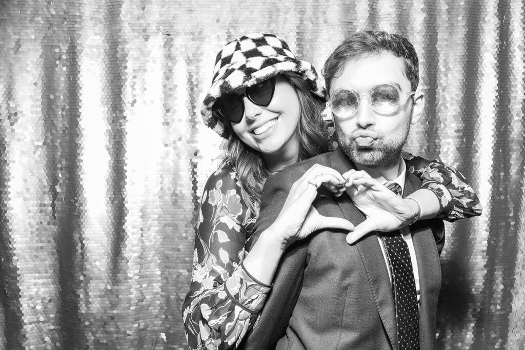 2 guests wearing hats and props doing a heart shape with her hand, posing against a silver sequins backdrop during a festive party entertainment