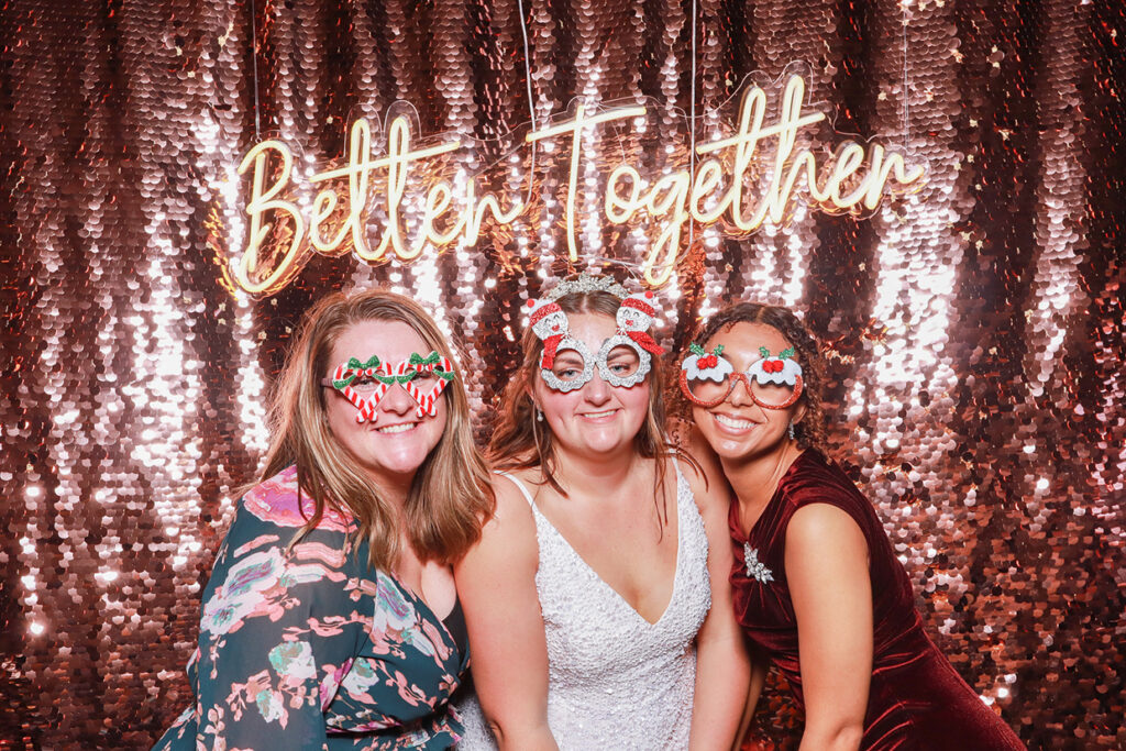 3 guests wearing Christmas props posing against a champagne sequins backdrop and a Better Together neon sign, during a photo booth party rental 