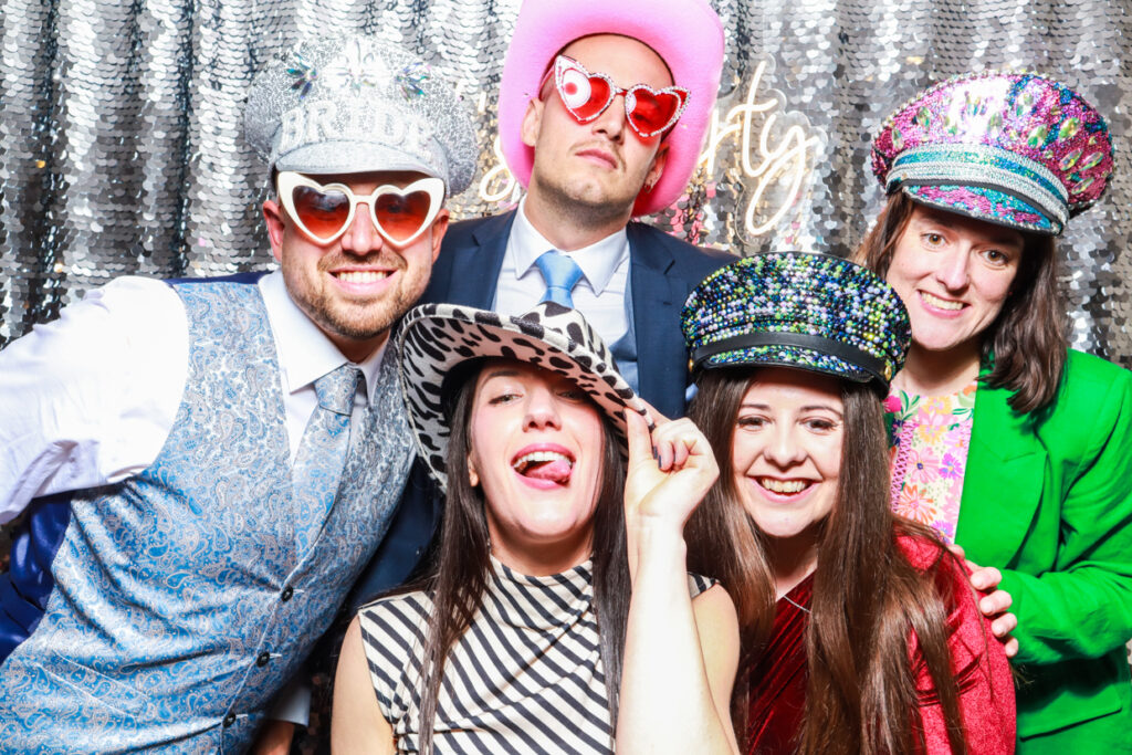 a large group of guests wearing fun props, posing against a shiny silver sequins backdrop with a lets party neon sign during a wedding party entertainment