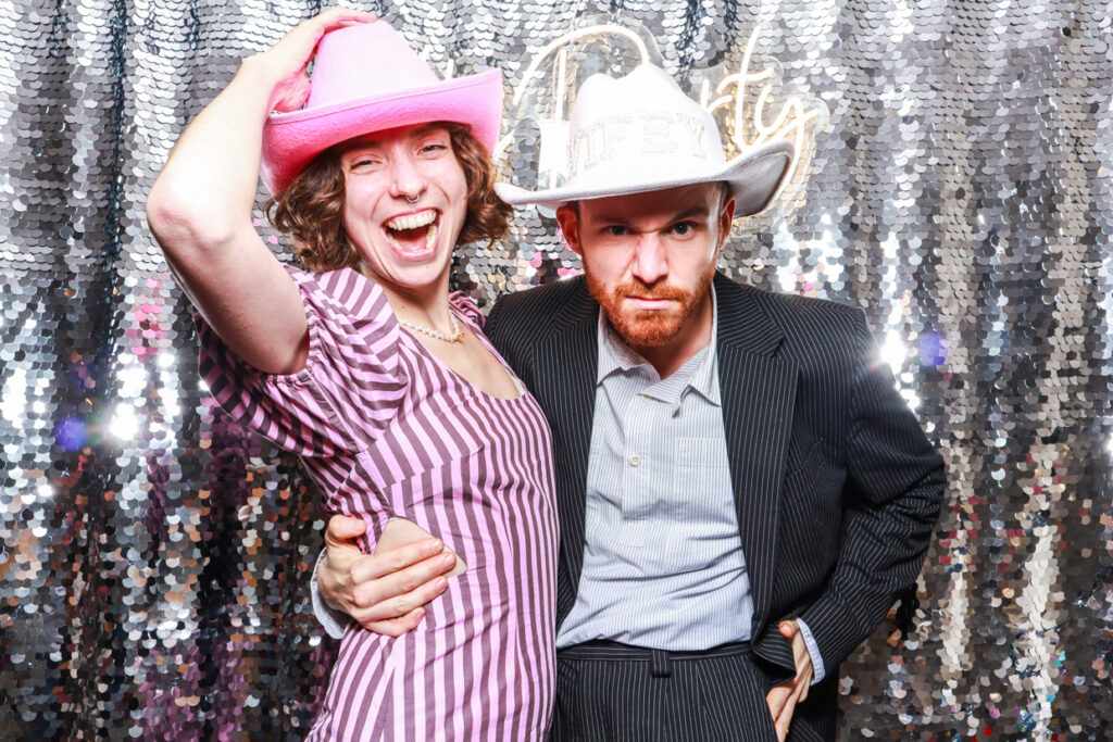 a couple being really funny and wearing cowboy hats, posing against a shiny silver sequins backdrop with a lets party neon sign during a wedding party entertainment