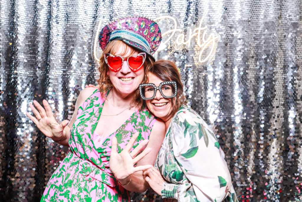 2 ladies wearing fun props, posing against a shiny silver sequins backdrop with a lets party neon sign during a wedding party entertainment