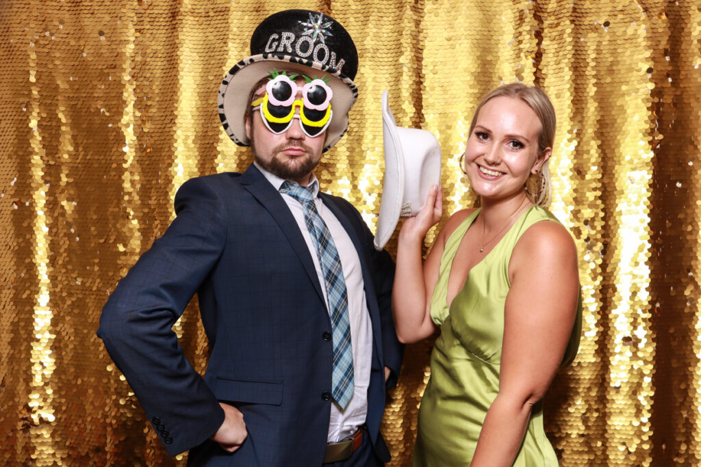 couple posing with fun props against a god sequins backdrop, during a party entertainment for a wedding party